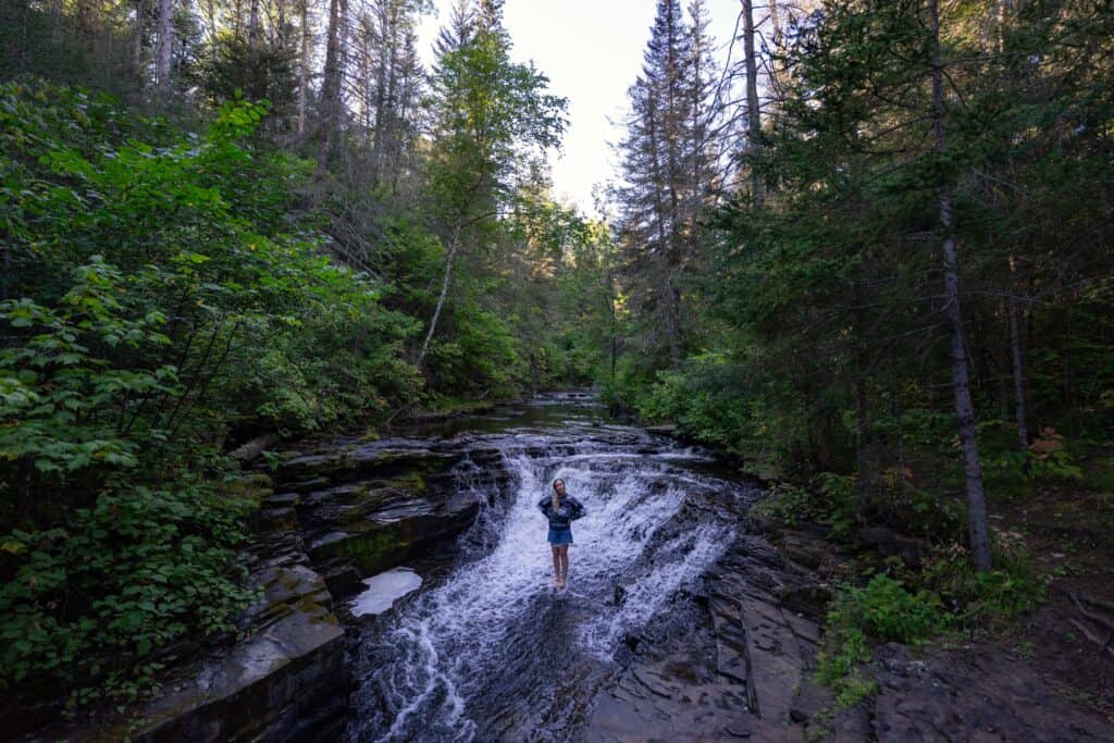 Person standing in a small waterfall along Cedar Creek at Cedar Falls Conservation Area near Thunder Bay, surrounded by forest.