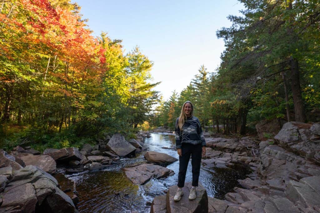 Blonde woman standing on rocks beside Duchesnay Creek, surrounded by colourful fall forest near North Bay, Ontario.