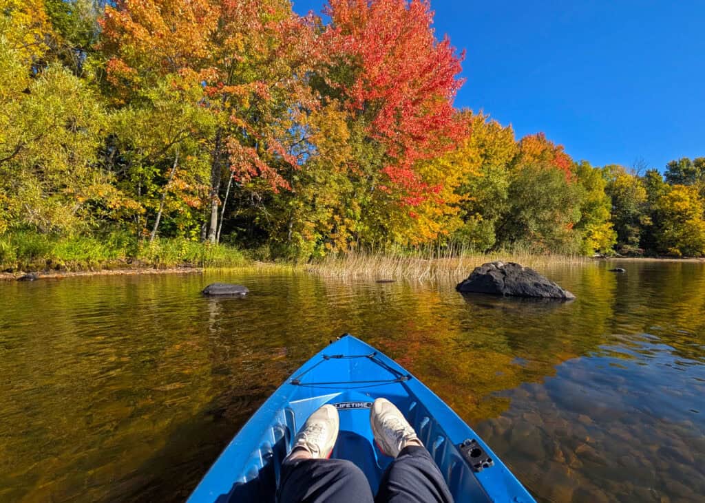 View from a blue kayak on Lake Nipissing, looking toward a shoreline covered in bright red, orange and yellow fall foliage in North Bay, Ontario.