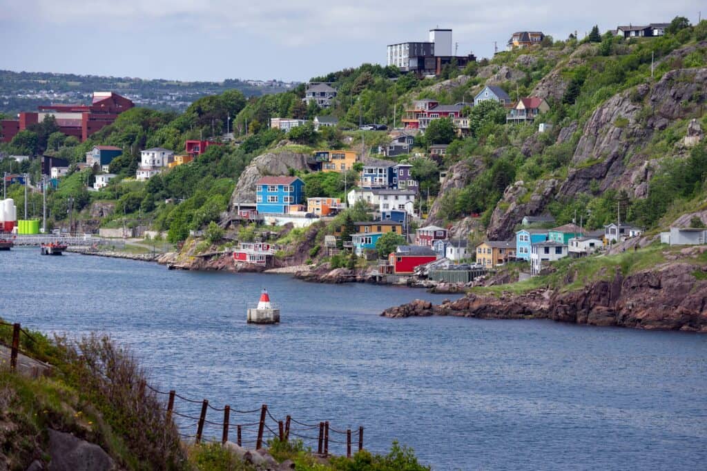 View across St. John’s Harbour toward the colourful houses of The Battery, built along the rocky slopes below Signal Hill in St. John’s, Newfoundland and Labrador, with a navigation marker in the water.